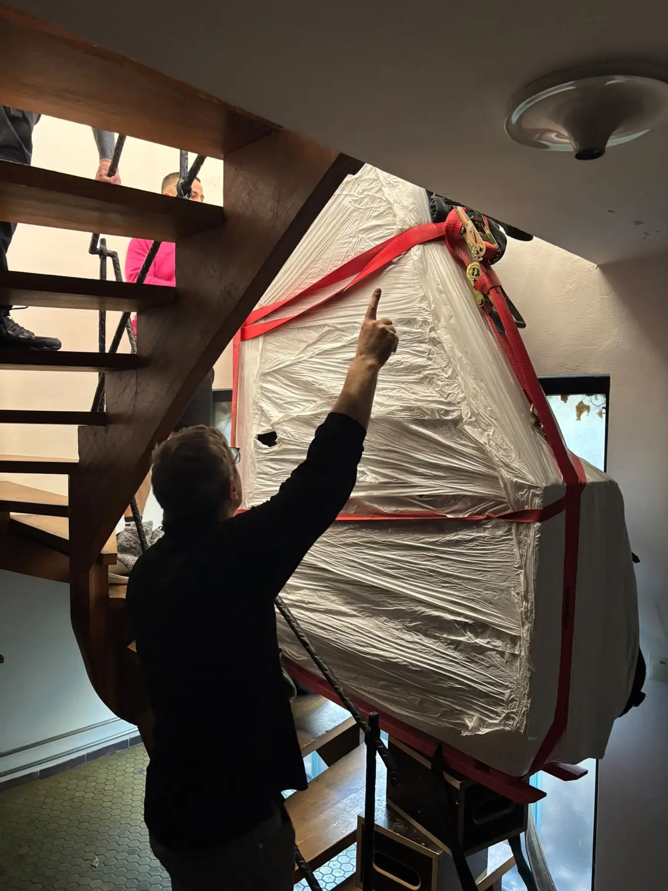 Ein verpackter Flügel wird über eine offene Holztreppe manövriert, ein Mitarbeiter dirigiert