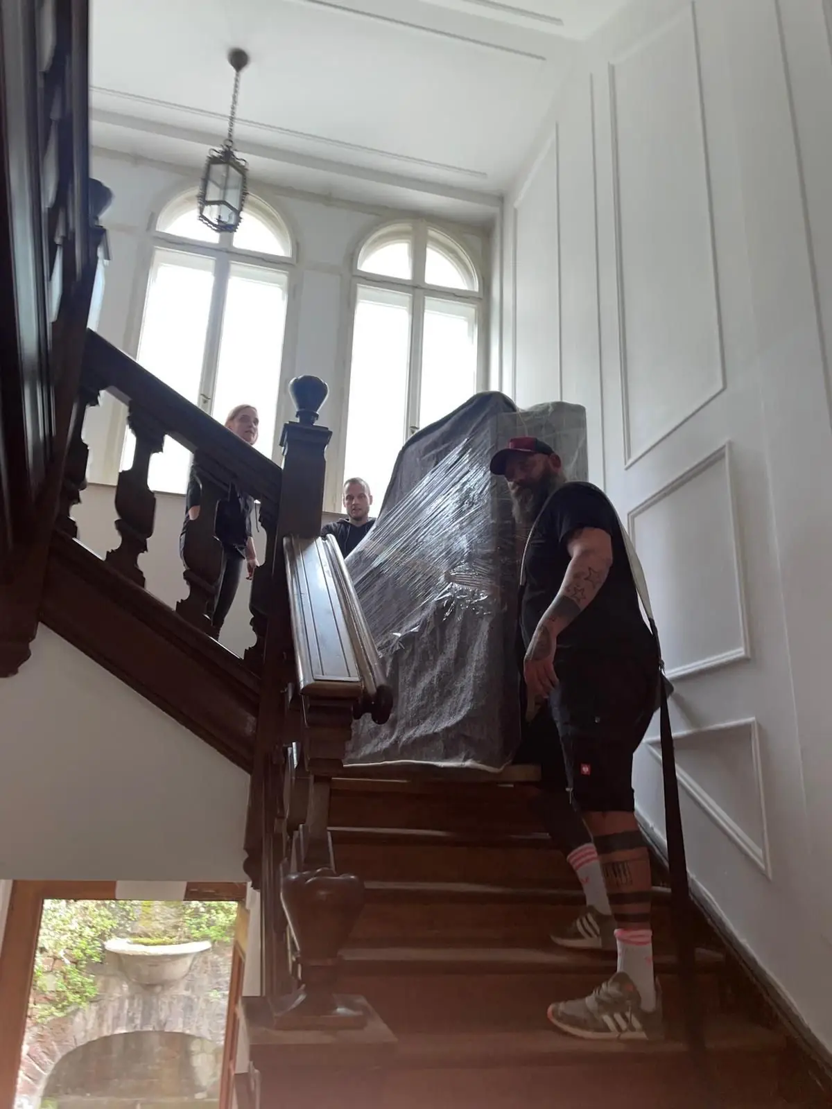 Ein verpacktes Klavier wird von zwei Mitarbeitern über eine geschwungene Altbautreppe nach oben getragen