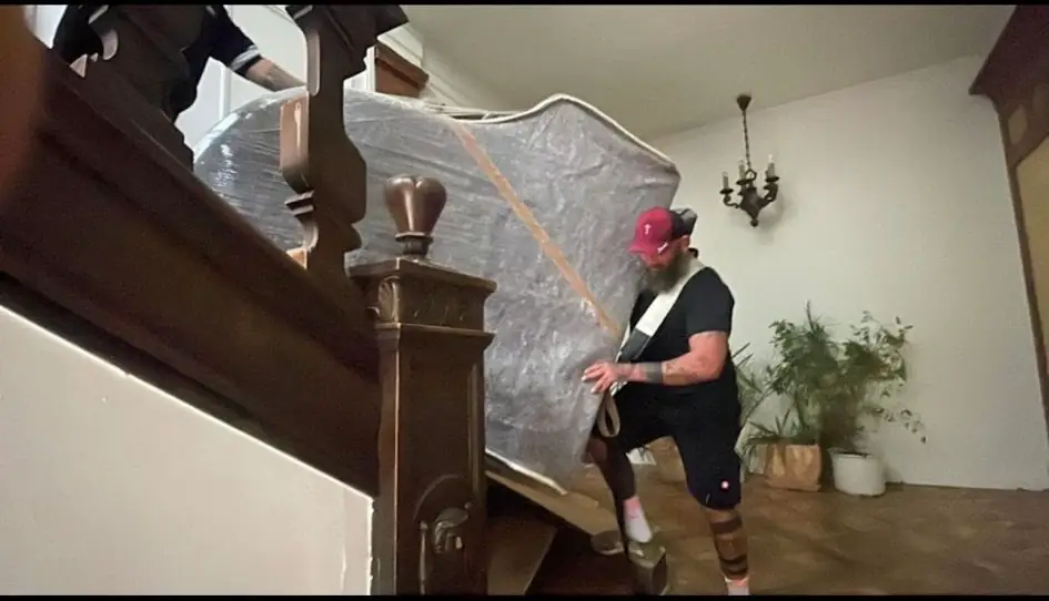 Ein Mitarbeiter führt ein gepolstertes Klavier kontrolliert eine Holztreppe hinunter