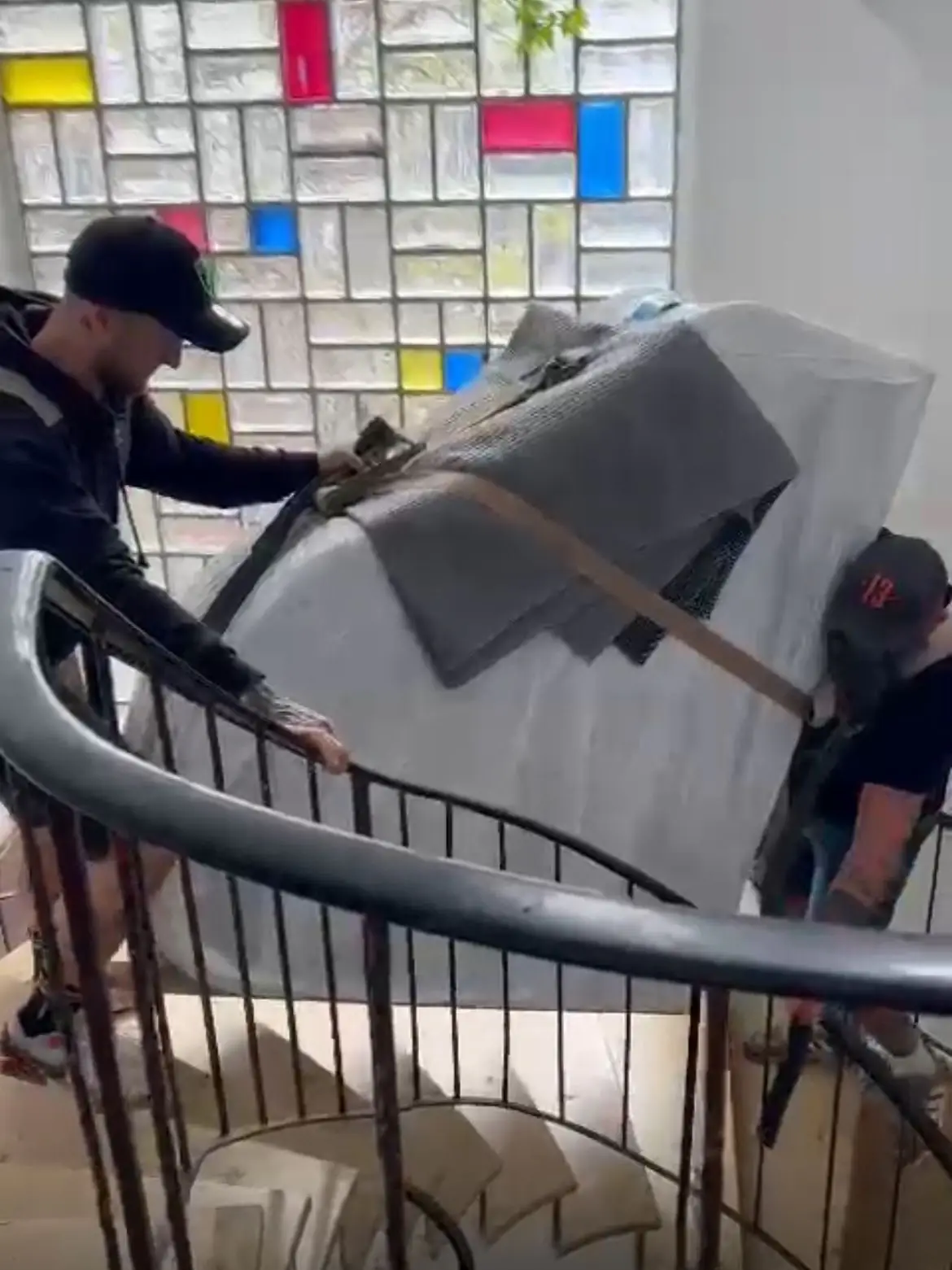 Zwei Mitarbeiter manövrieren ein gepolstertes Instrument über eine enge Wendeltreppe